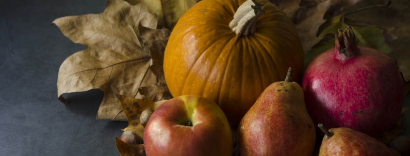 A flat lay of a pumpkin, a pomegranate, two pears and an apple on top of fall leaves