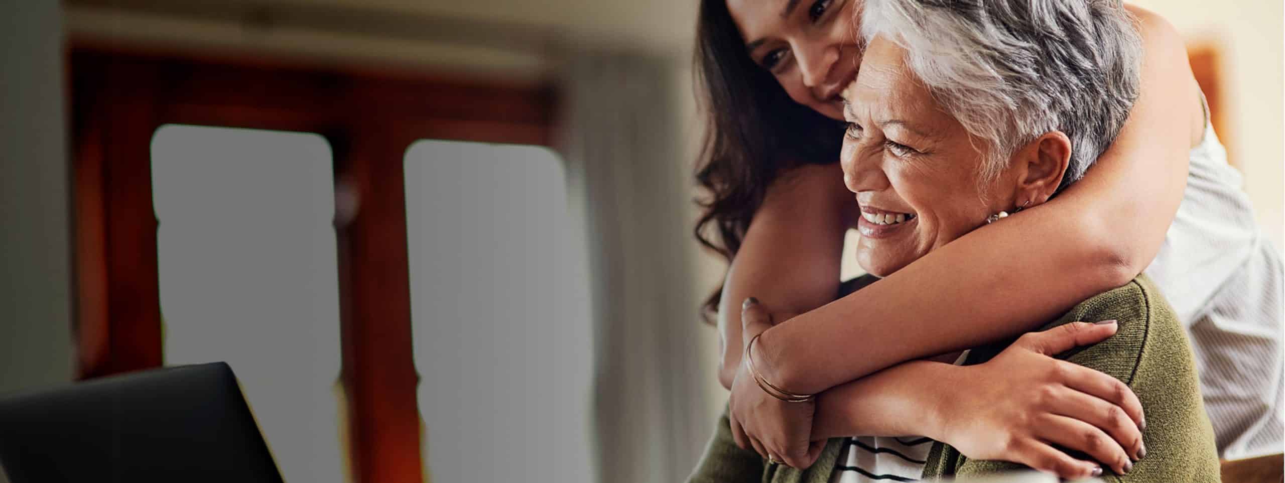 An elderly woman using her computer while her daughter hugs her from behind