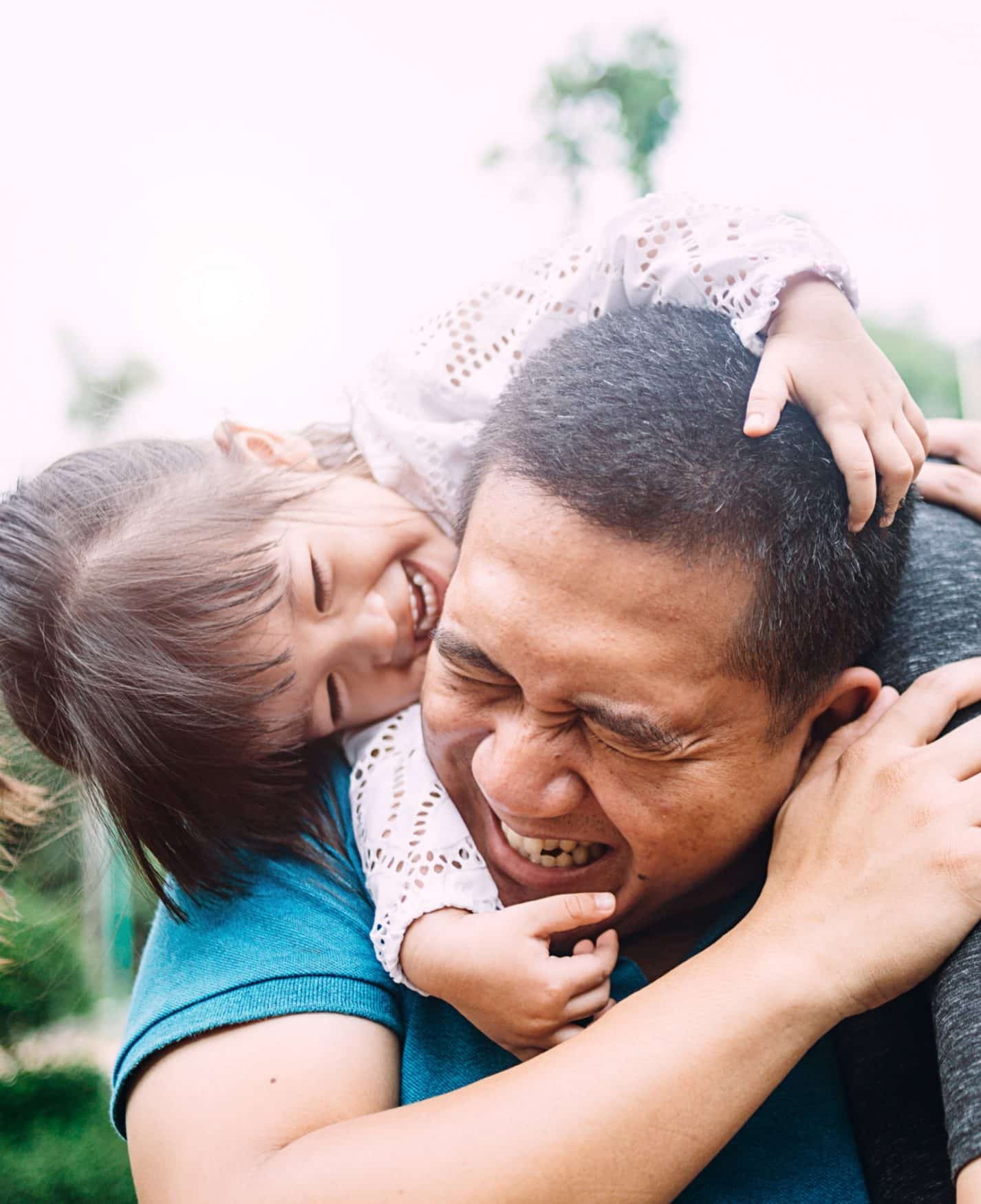 A dad and daughter laughing while he gives her a piggy back ride outside