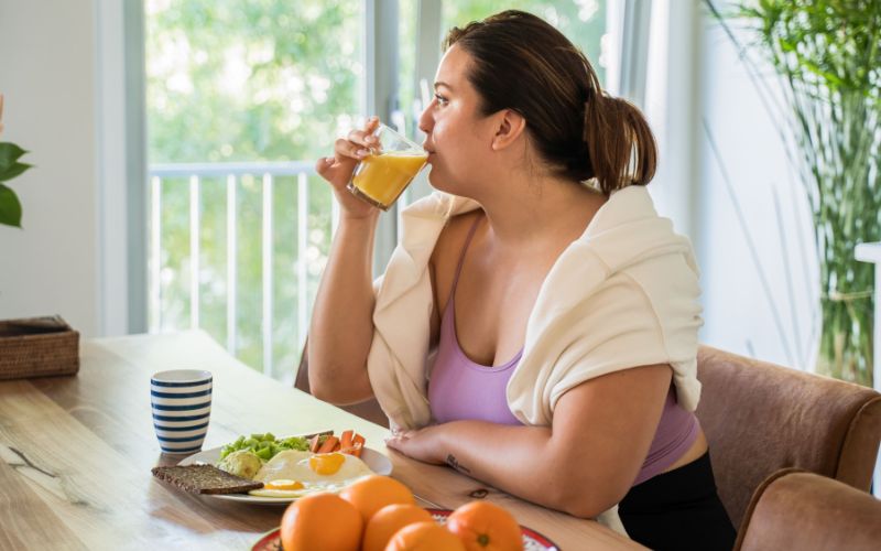 An overweight woman sits before a healthy meal sipping fresh juice.