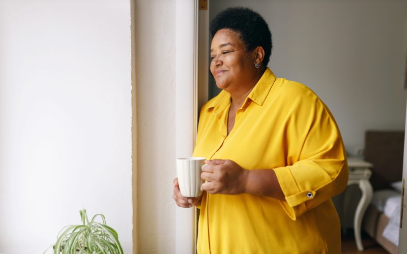 An African American woman smiles as she looks out the window holding a mug of tea.